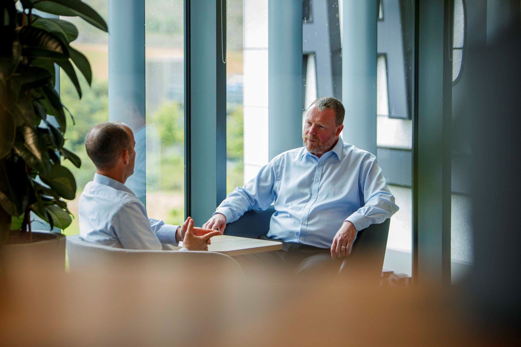 An image of two AAB employees sitting in the office wearing blue shirts and sharing coversation. An image of two AAB employees sitting in the office wearing blue shirts and sharing coversation.