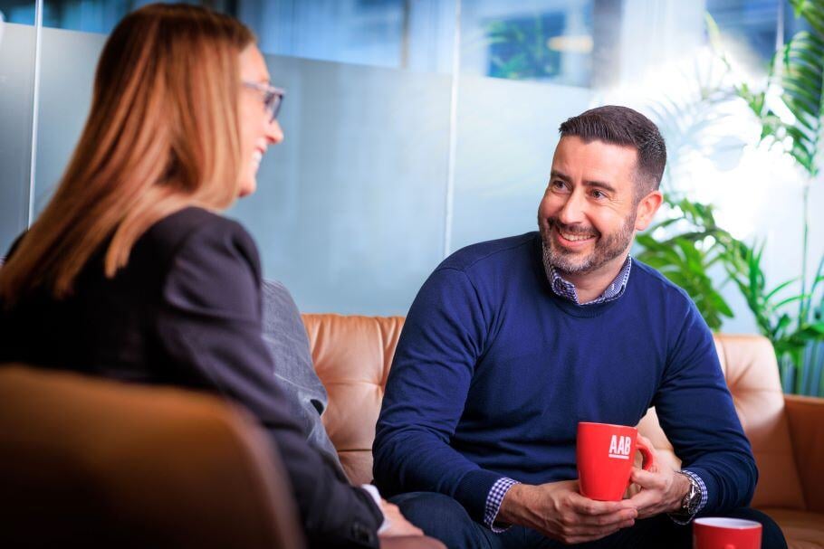 An image of two AAB employees sitting on a couch in conversation. One of them is smiling and carrying a red AAB branded mug.