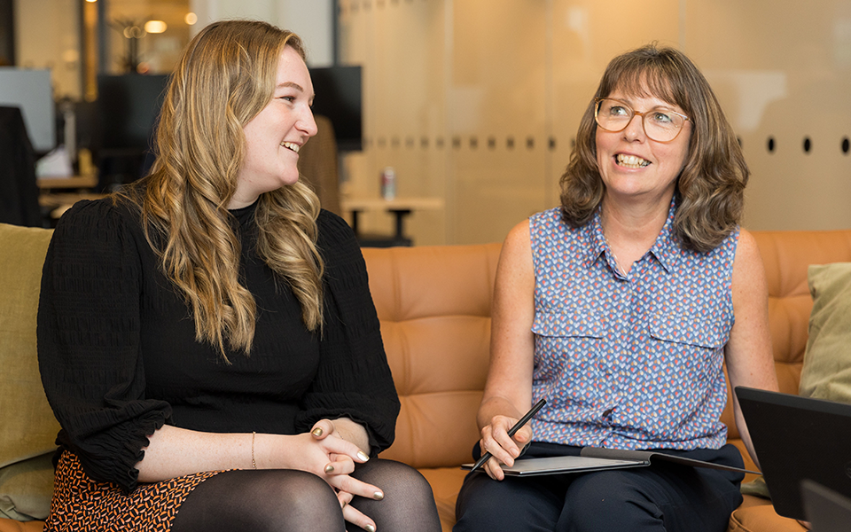 An image of two AAB employees sitting in the office with a notebook and sharing conversation. An image of two AAB employees sitting in the office with a notebook and sharing conversation.