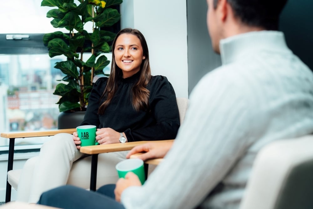 An image of Daniel Lightbody, Lori Young sitting in the office having a conversation with a cup of coffee.