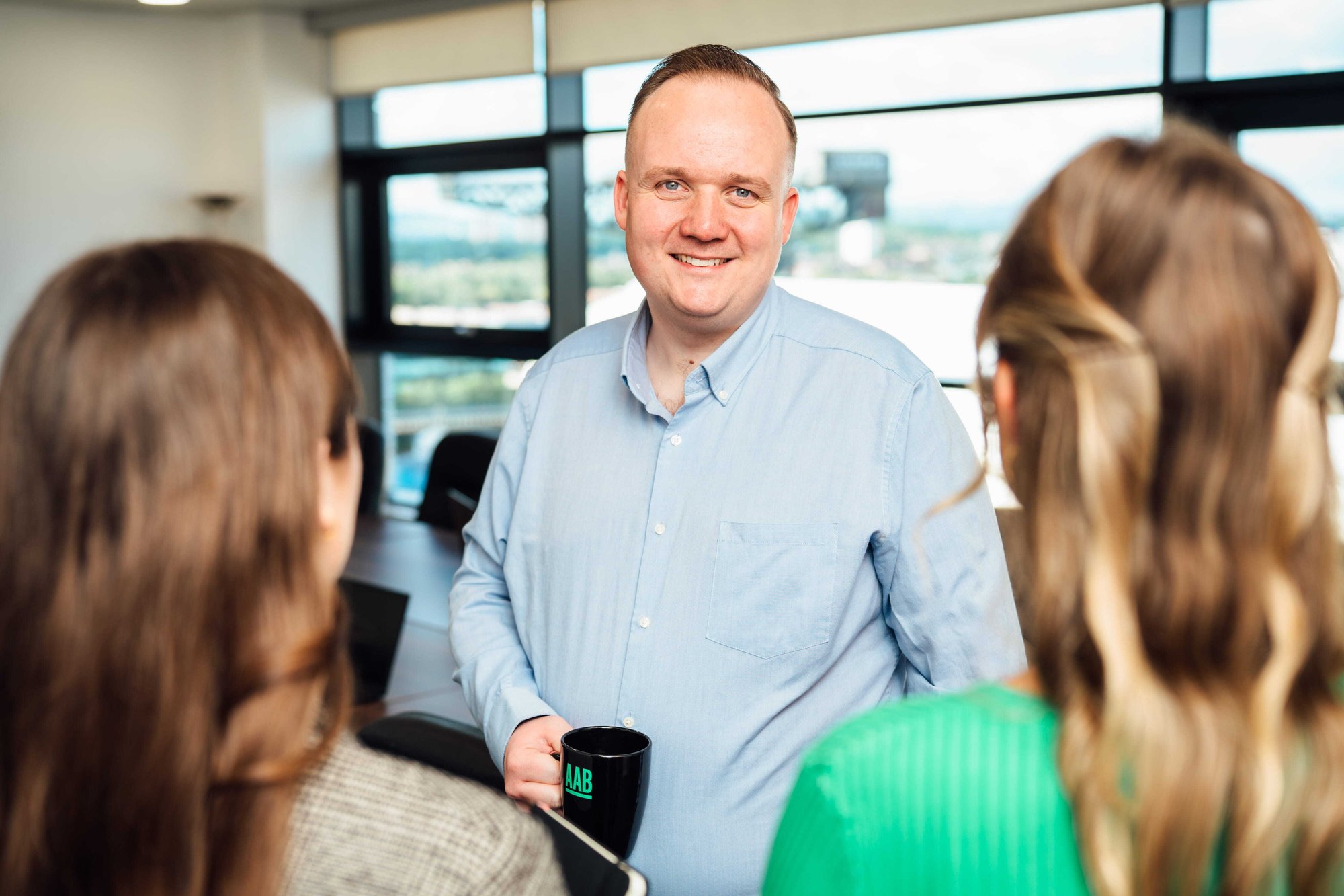 An image of Josh McCranor wearing a blue shirt and smiling at the camera. He is holding an AAB branded mug.