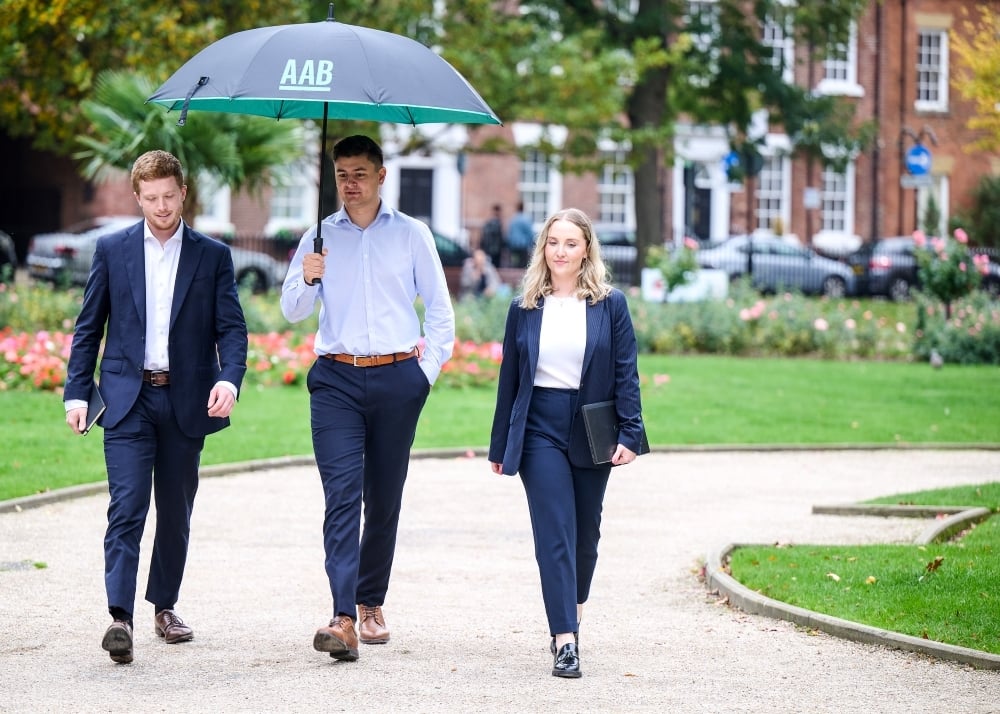 An image of three AAB employees holding an AAB branded umbrella and walking through a park.