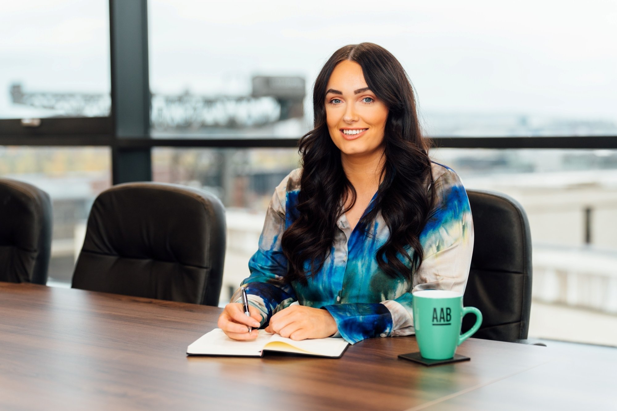 An image of Olivia Russell wearing a blue shirt and sitting at a desk writing in her notebook. She also has an AAB branded mug. An image of Olivia Russell wearing a blue shirt and sitting at a desk writing in her notebook. She also has an AAB branded mug.