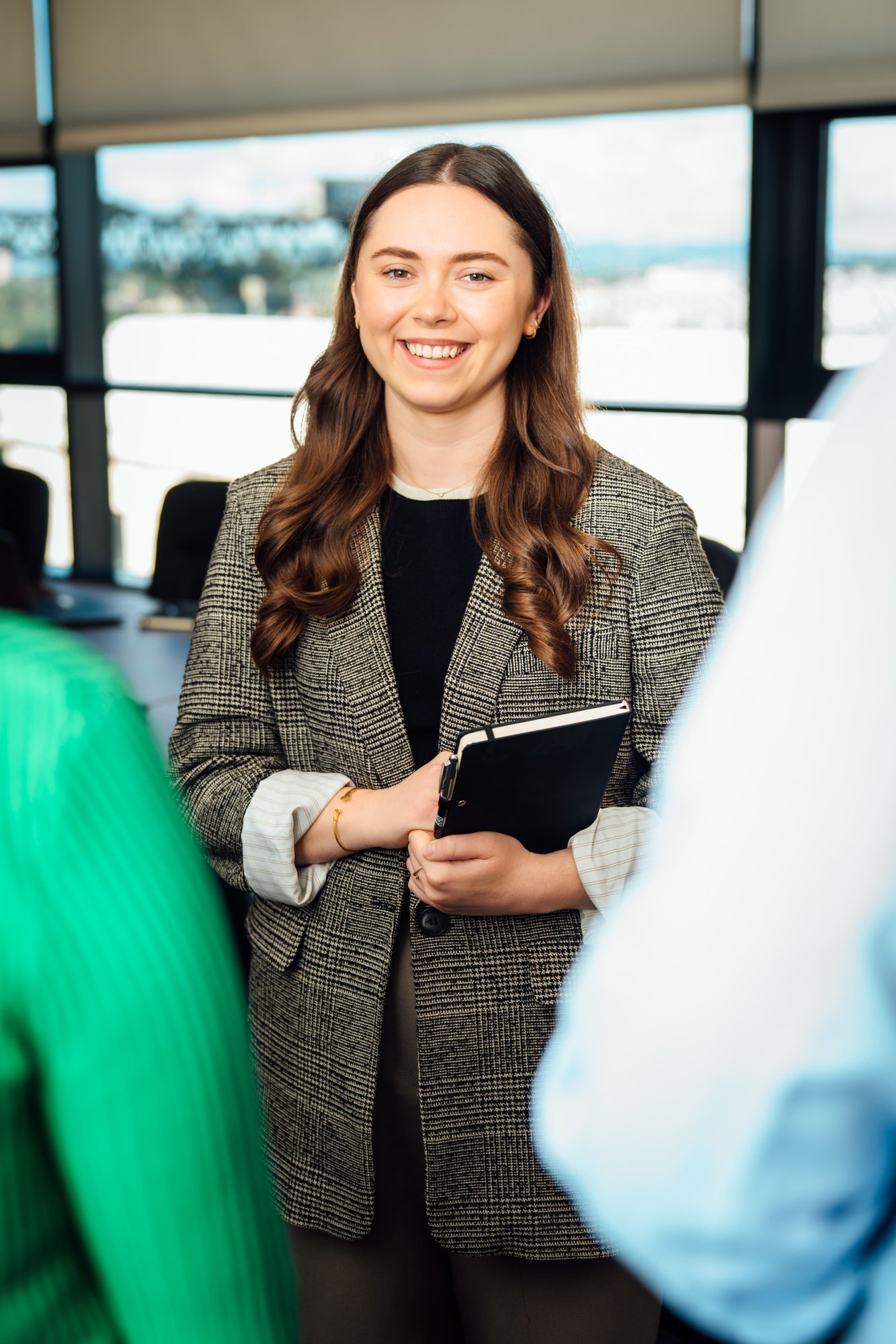 An image of Zoe Dollard wearing a brown blazer and black top. She is holding an AAB notebook and smiling at the camera.